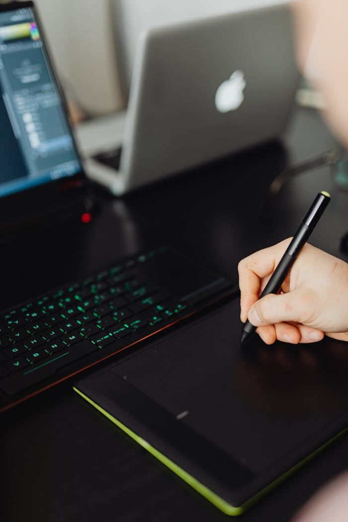 Hand holding stylus pen writing on digital tablet with laptop setup, perfect for creative workspaces.